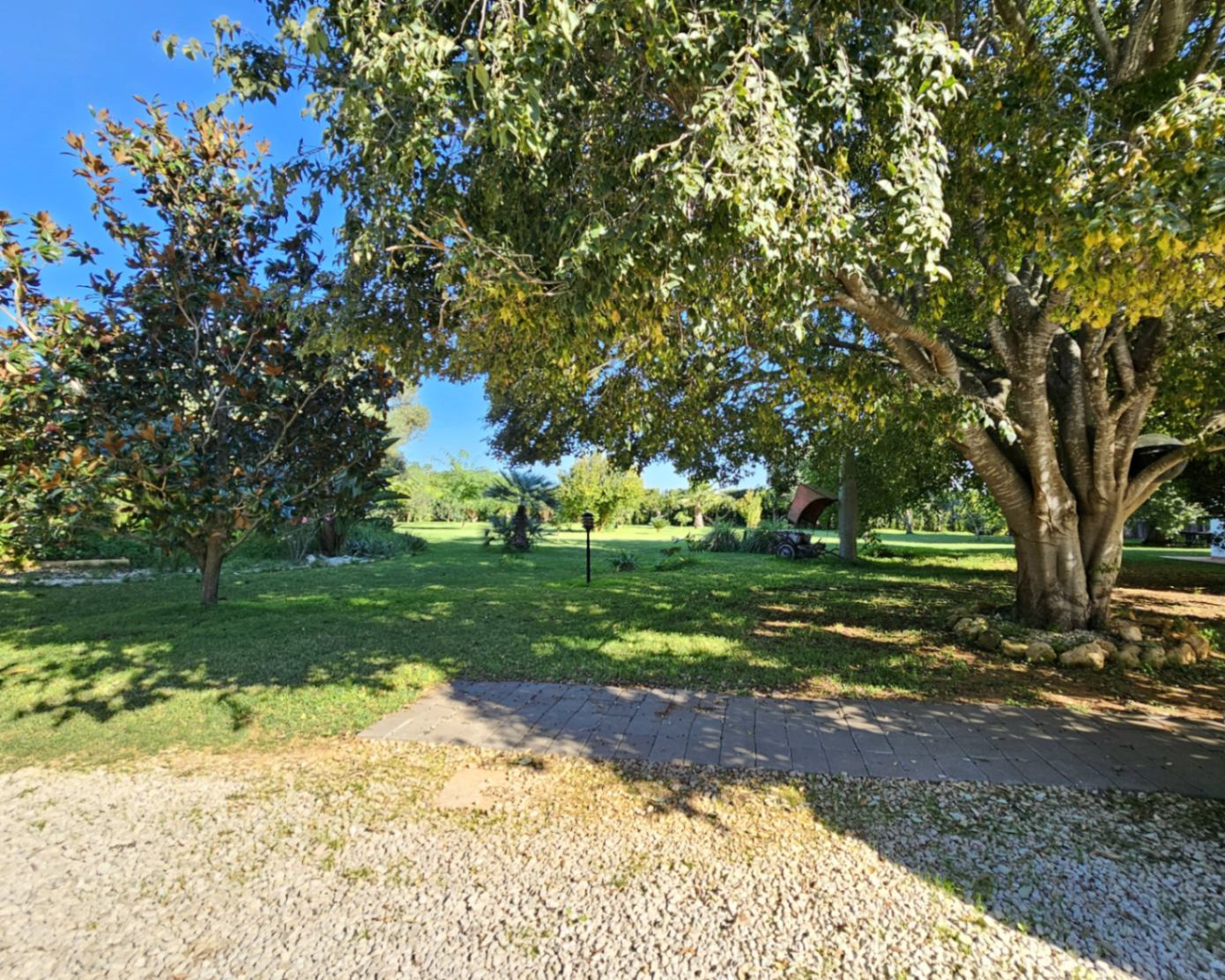 Verzorgde mediterrane tuin met volwassen schaduwbomen, uitgestrekte gazons en paden van grind en natuursteen op het perceel.