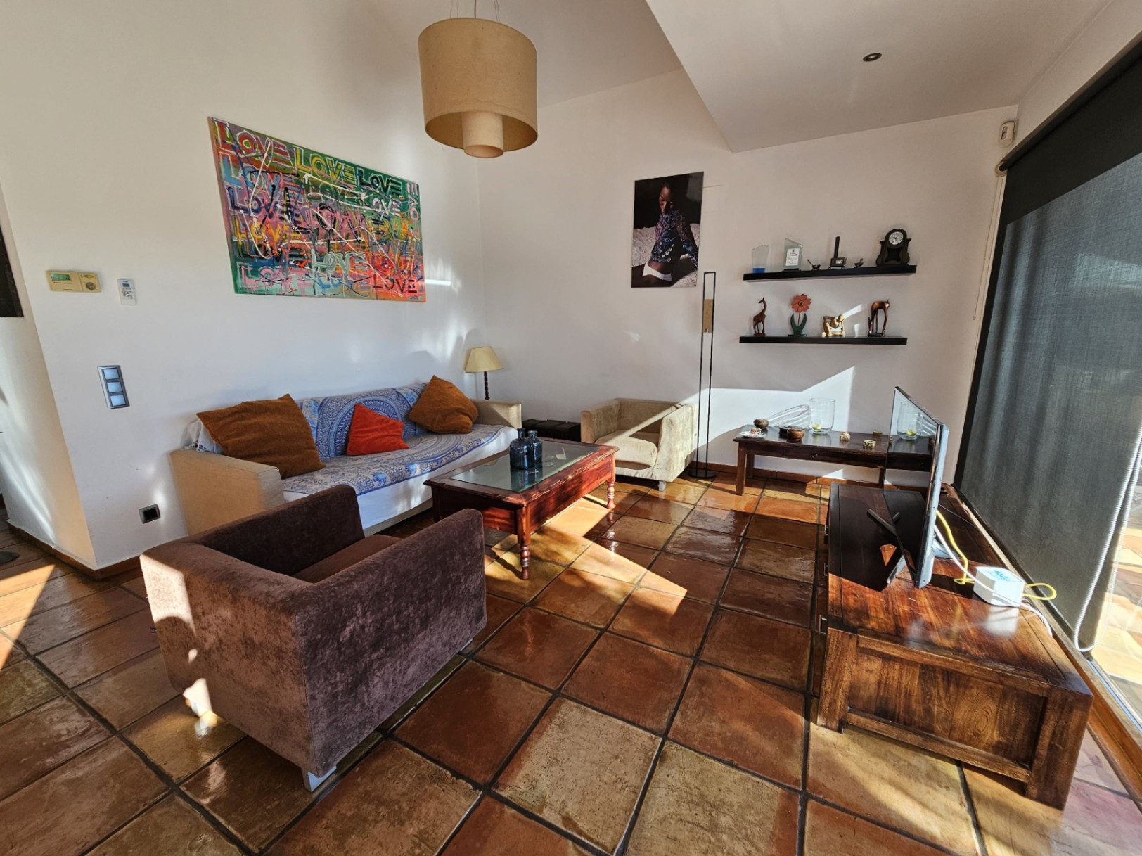 Woonkamer met handgemaakte terracotta vloeren en houten meubels. Natuurlijk licht verlicht de kleurrijke kunstwerken en de rustieke afwerking van de ruimte.