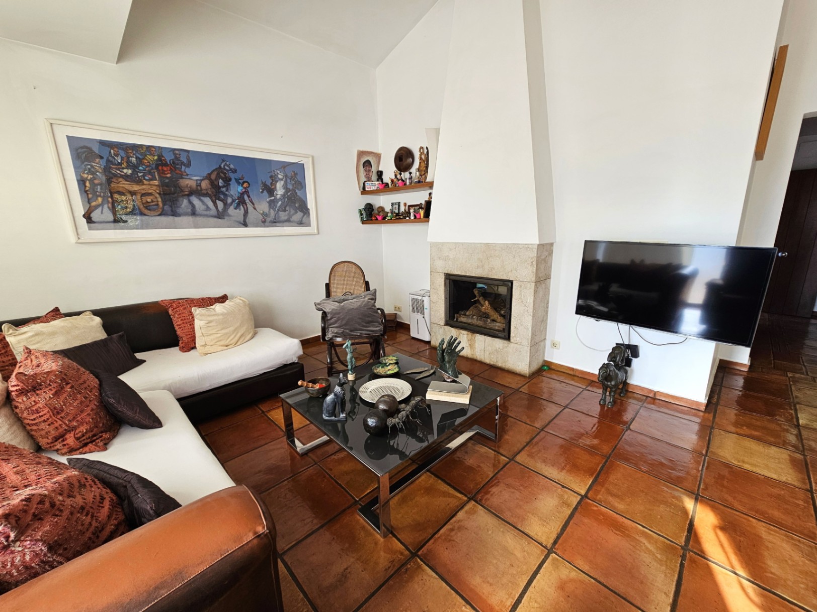 Woonkamer met handgemaakte terracotta vloertegels en een ingebouwde stenen open haard. De ruimte heeft hoge plafonds, modern meubilair en veel lichtinval.