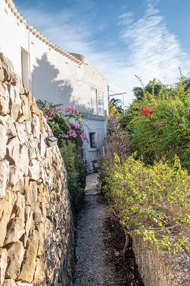 Buitenpad omzoomd door traditionele natuurstenen muren en mediterrane beplanting, lopend langs de witgestuukte gevel van de villa.