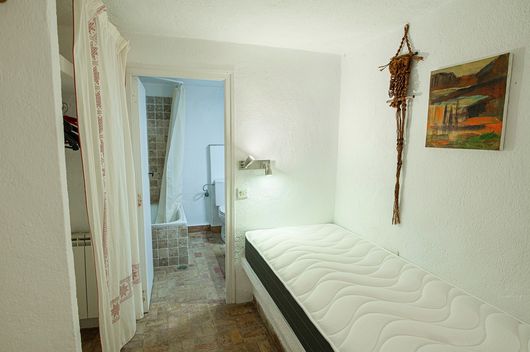Functionele slaapkamer met eenpersoonsbed op een vaste basis, witte muren met textuur en een eigen badkamer met rustieke tegels in aardetinten.