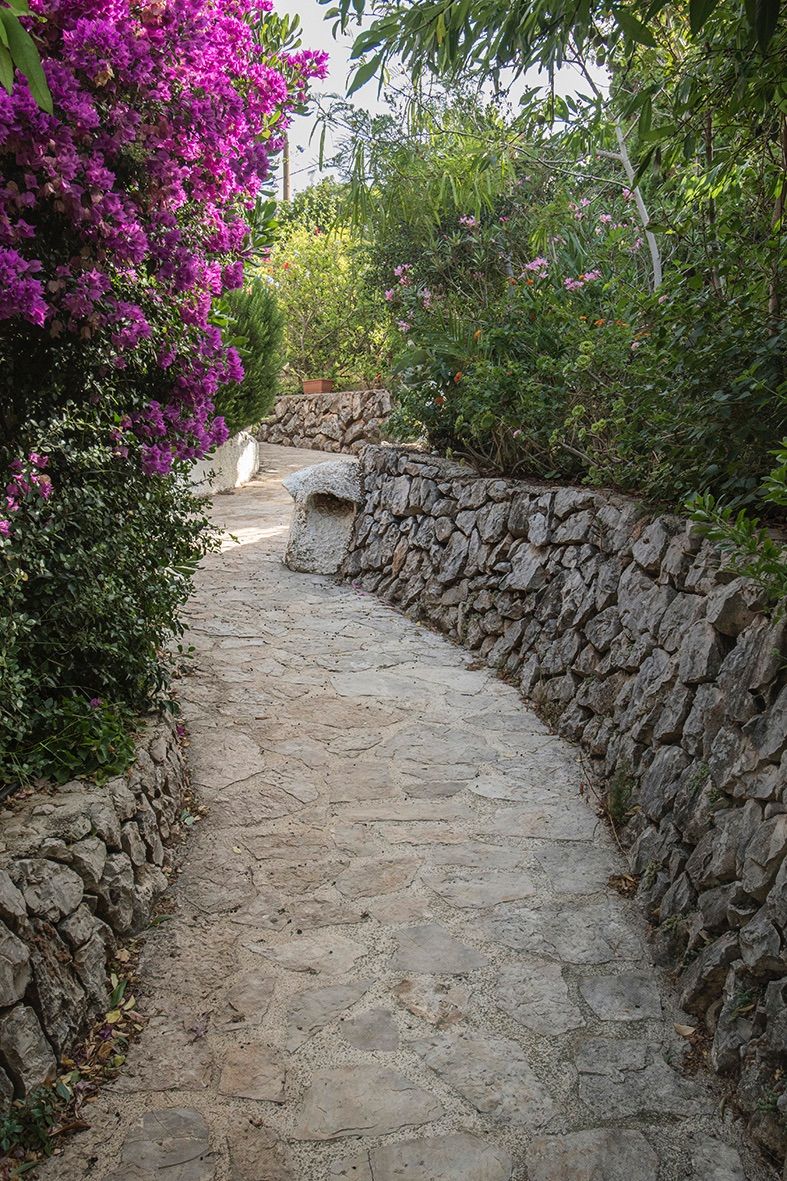 Natuurstenen tuinpad omzoomd door traditionele droge stenen muren en weelderige mediterrane beplanting met paarse bougainvillea.