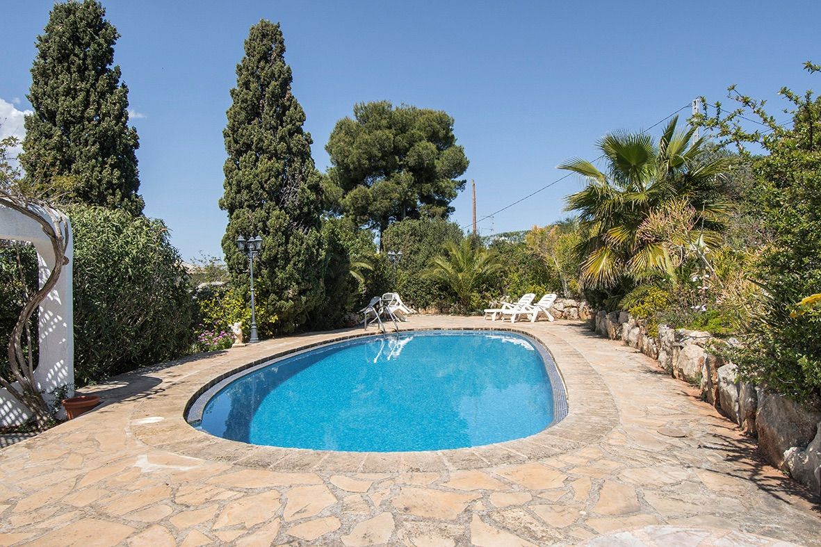 Ovaalvormig zwembad met een terras van natuursteen, omgeven door een mediterrane tuin met hoge cypressen en palmbomen.