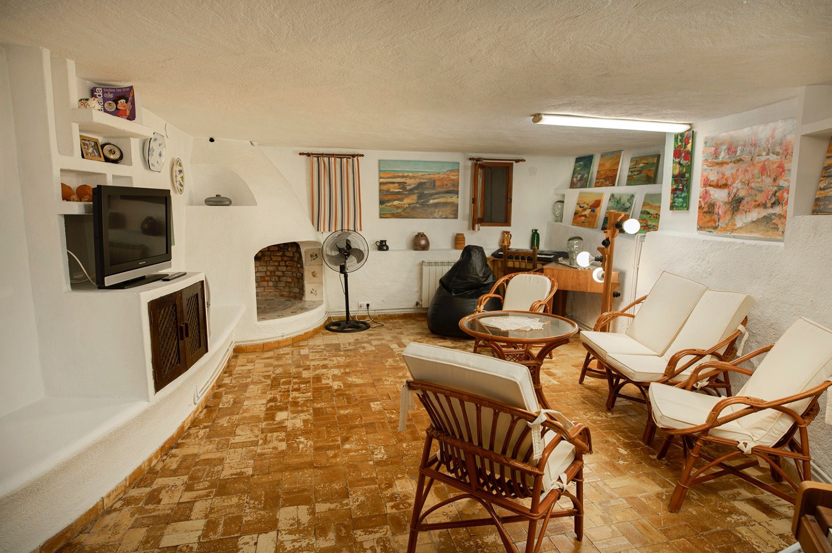 Rustieke woonkamer met witgepleisterde muren, een ronde open haard, terracotta tegelvloer en bamboe meubilair met witte kussens.