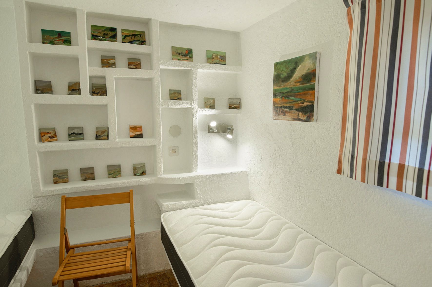 Slaapkamer met witte muren en ingebouwde nissen voor kunstwerken, voorzien van een eenpersoonsmatras, een houten klapstoel en wandverlichting.
