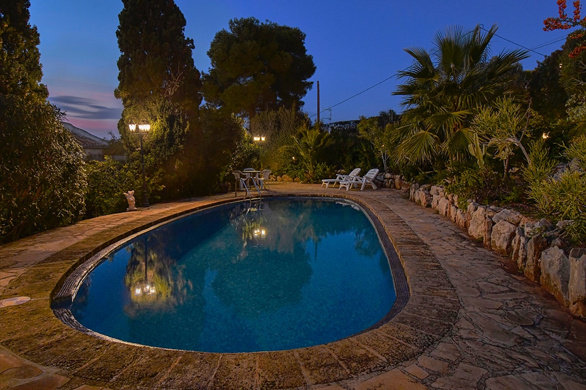 Verlicht buitenzwembad bij schemering, omgeven door natuurstenen bestrating en een mediterrane tuin met palmbomen en cipressen.