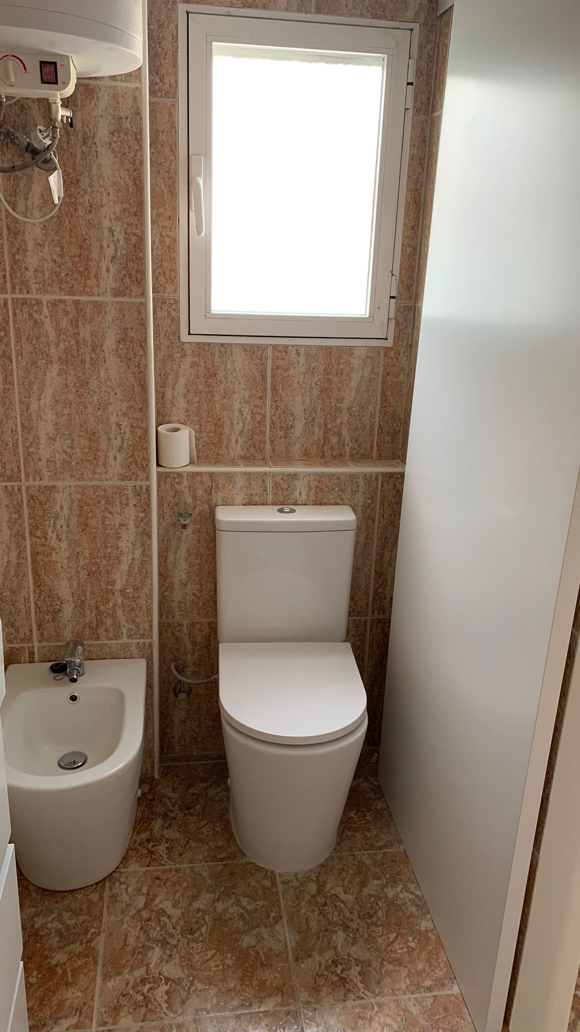 Badkamer met wit porseleinen toilet en bidet, aardetinten tegels met marmerlook en een raam voor natuurlijke lichtinval en ventilatie.