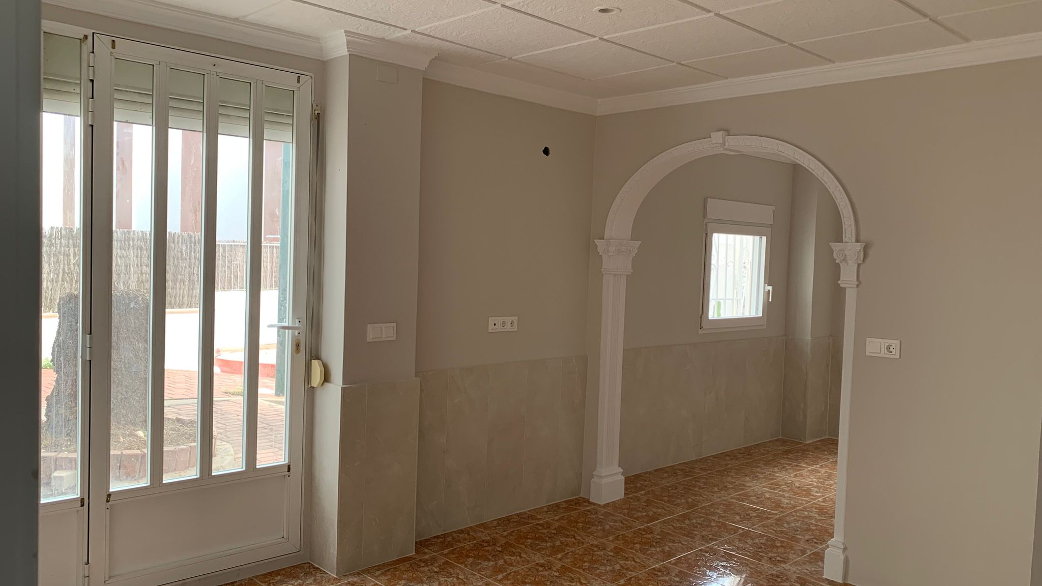 Woonkamer met keramische tegelvloer, betegelde lambrisering en een decoratieve witte boog. Een glazen deur biedt directe toegang tot de buitenruimte.