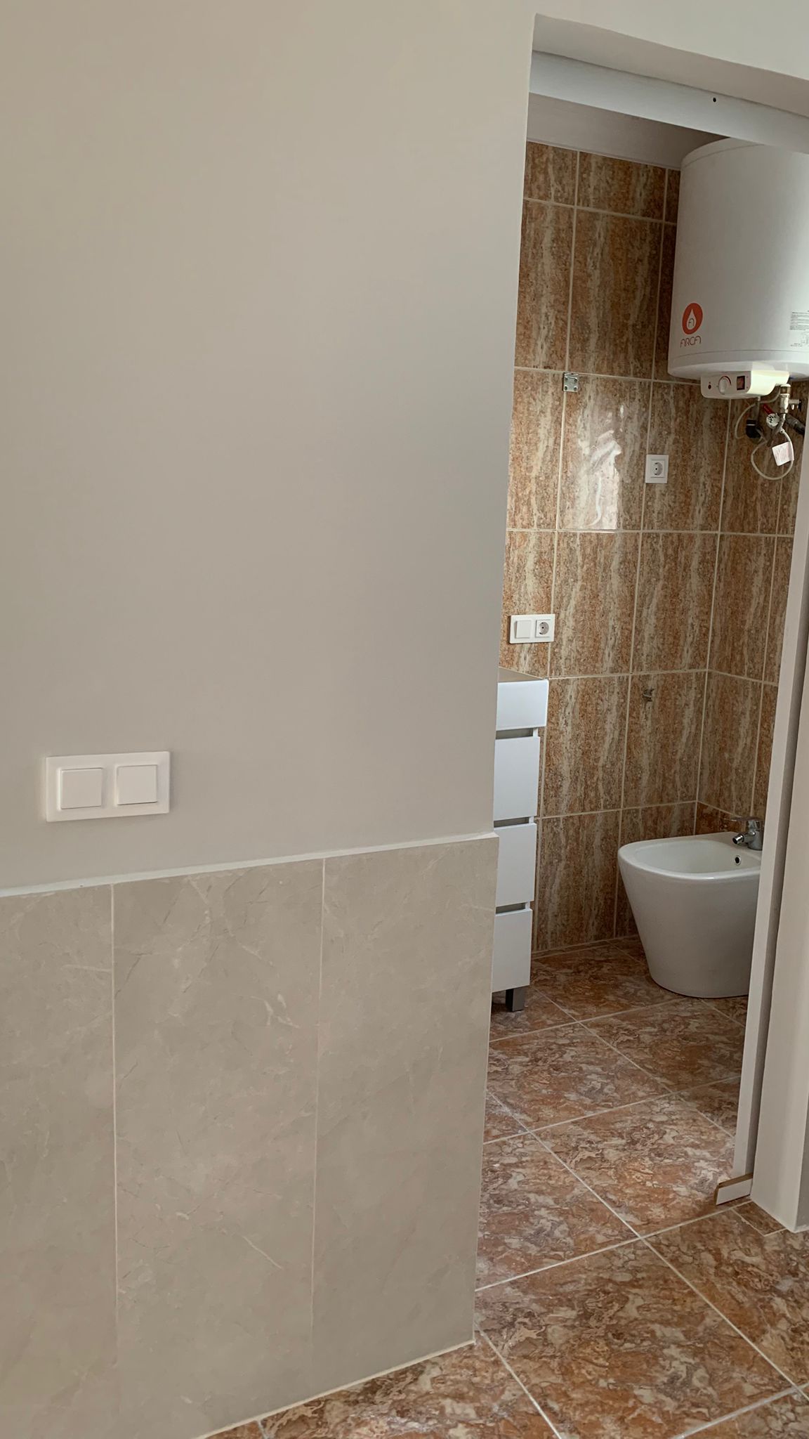 Zicht op de badkamer met bidet, wit opbergmeubel en elektrische boiler, afgewerkt met bruine gemarmerde wand- en vloertegels.
