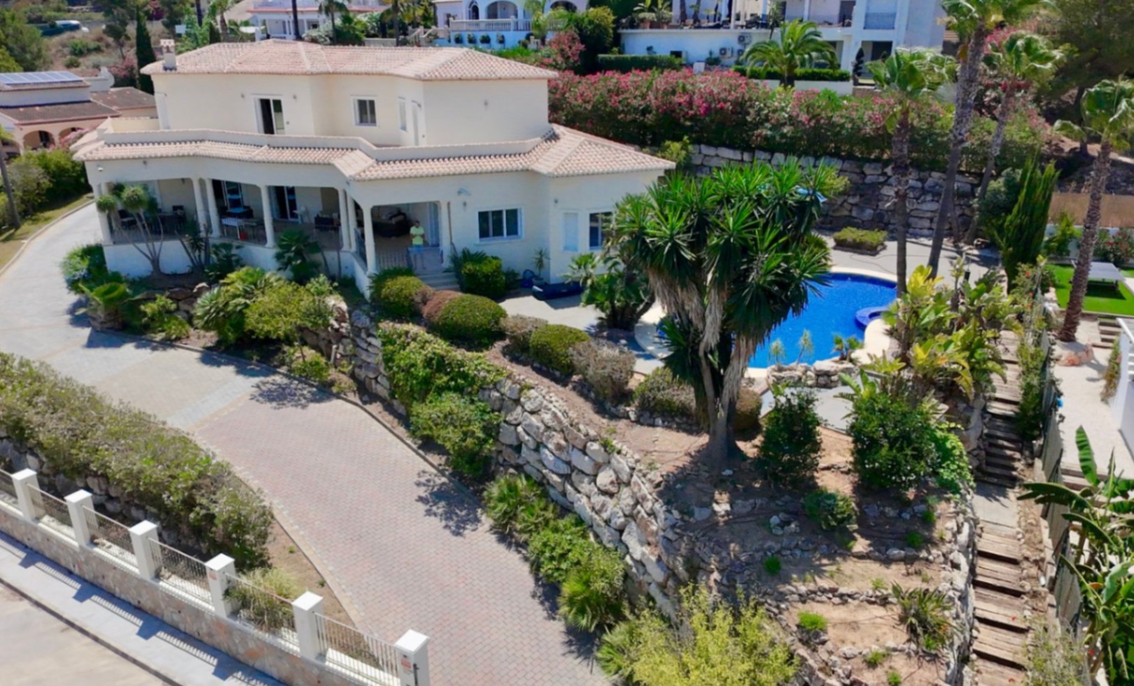 Buitenaanzicht van de villa met witte gevel, pannendaken, ruime geplaveide oprit, natuurstenen muren en aangelegde mediterrane tuinen met een zwembad.