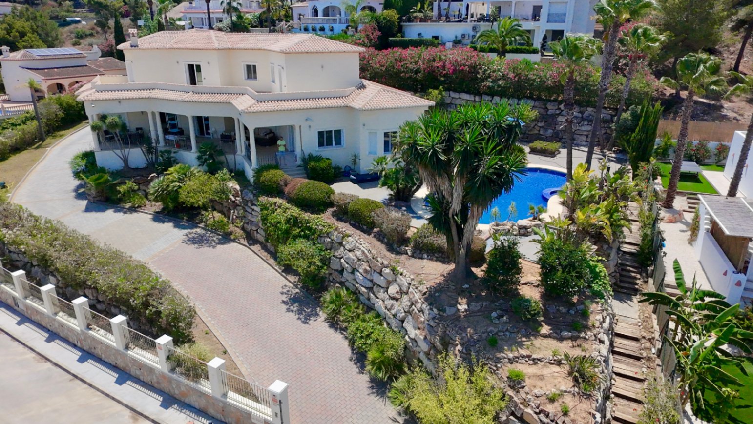 Luchtfoto van de villa met mediterrane architectuur, een privézwembad omgeven door palmbomen en natuurstenen muren op een verhoogd perceel in Piver, Jávea.
