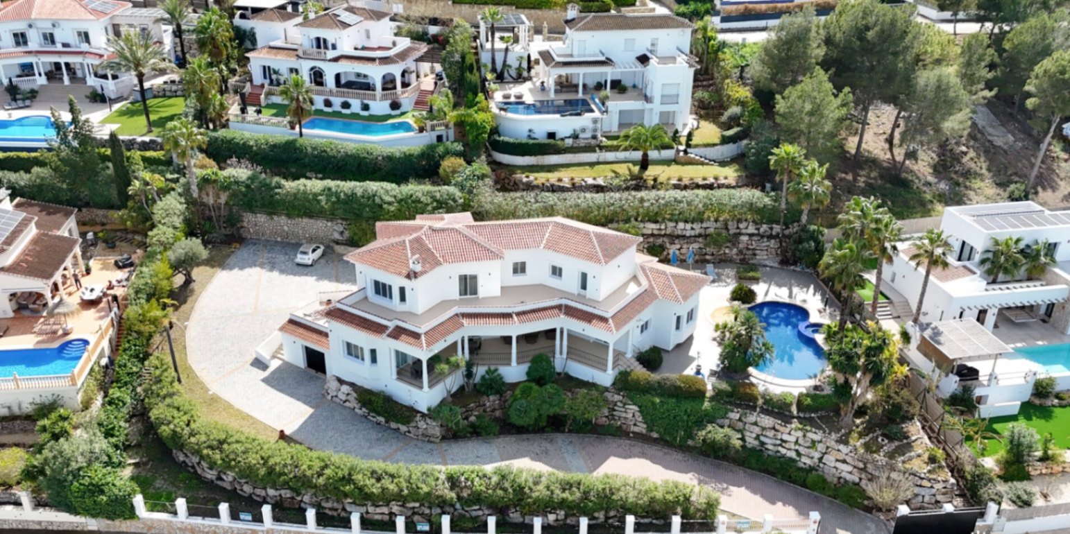 Luchtfoto van de villa met witte gevels en pannendak, voorzien van een ruime geplaveide oprit, natuurstenen muren en een privézwembad omgeven door palmbomen.