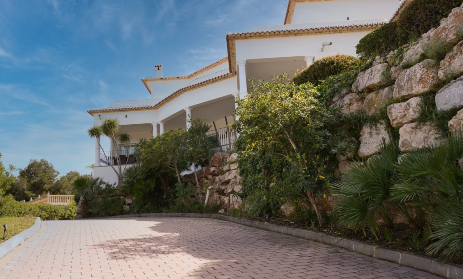 Mediterrane villa met witte kolommen, overdekte terrassen en een geplaveide oprit, geflankeerd door een natuurstenen muur en weelderige beplanting.