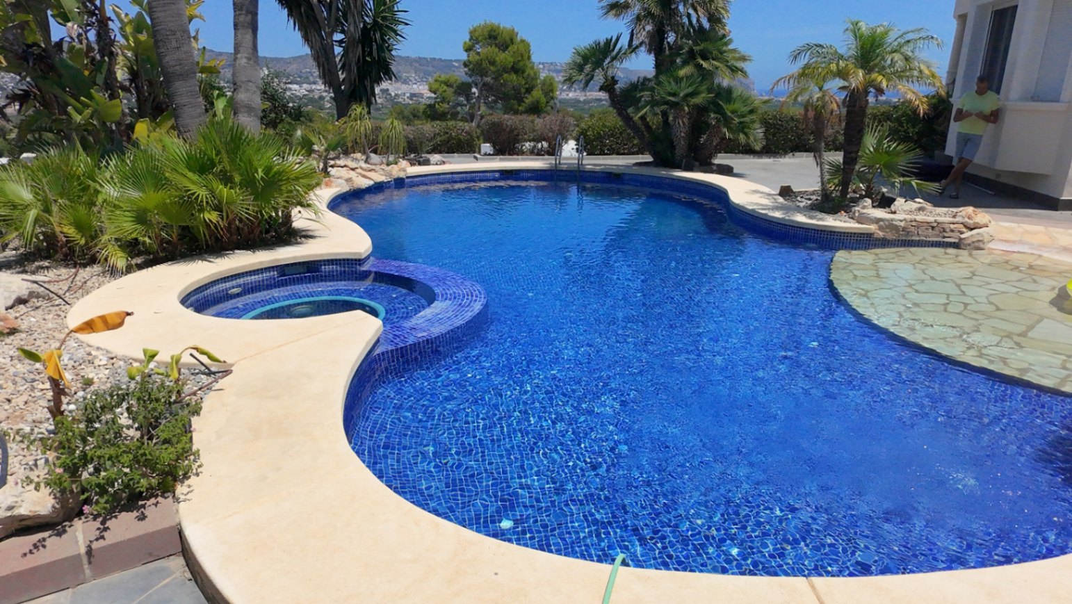 Organisch gevormd zwembad met blauwe mozaïektegels, geïntegreerde ronde spa en natuurstenen instap, omringd door palmbomen en mediterrane beplanting.
