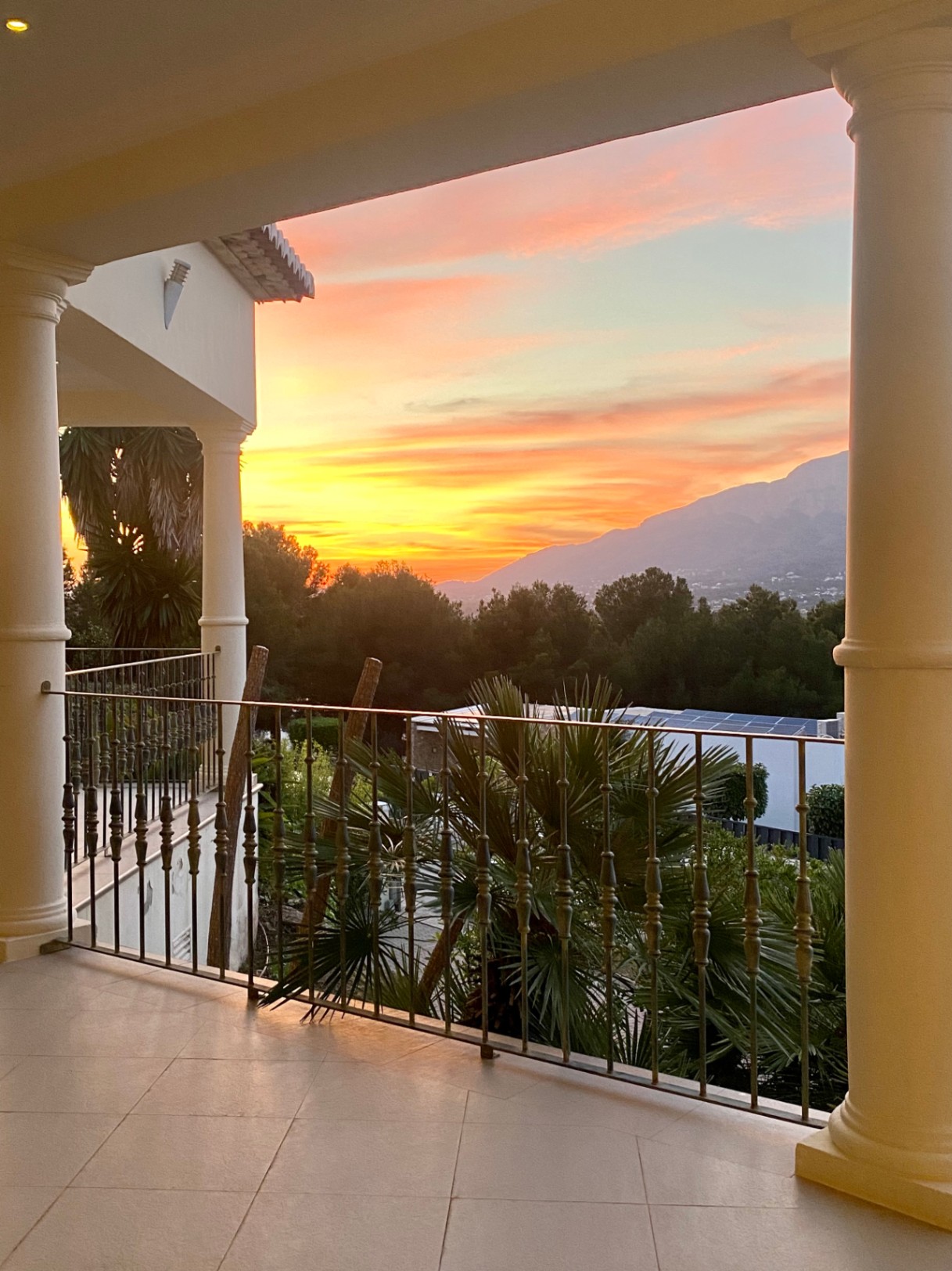 Overdekt terras met klassieke zuilen en smeedijzeren balustrade, uitkijkend op de contouren van de Montgó bij zonsondergang.
