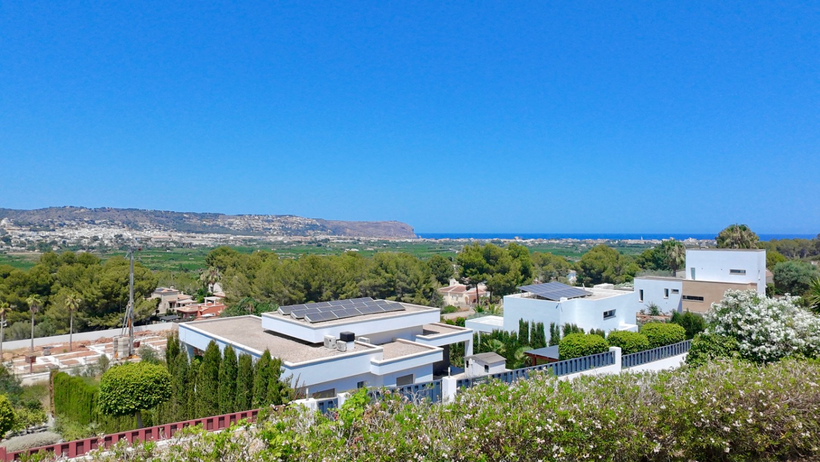 Verhoogd panoramisch uitzicht over de Middellandse Zee en Cabo de San Antonio, met moderne villa's voorzien van zonnepanelen in een groene, bosrijke omgeving.